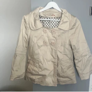 Beige trekvartsärmad kappa  - Snygg beige kappa med trekvartsärm och stora knappar framtill. Jackan har en klassisk krage och ett mönstrat foder som ger en extra touch. Perfekt för vår och höst!