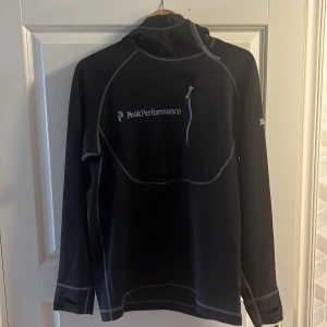 Svart tröja från Peak Performance - Säljer en svart tröja från Peak Performance i storlek M. Den har en dragkedja vid halsen och är perfekt för kyliga dagar. Tröjan är långärmad och har en sportig look med vita sömmar som detaljer. Perfekt för träning eller vardagsbruk!