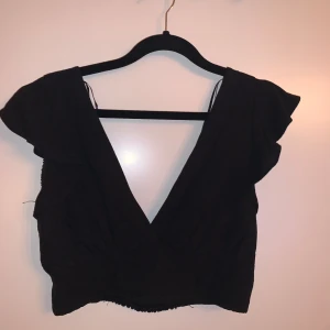 Svart kortärmad topp - Säljer en snygg svart topp med korta ärmar och en djup v-ringning. Toppen är croppad och har en stilren design. Från Pull&Bear i storlek M. Aldrig använd 