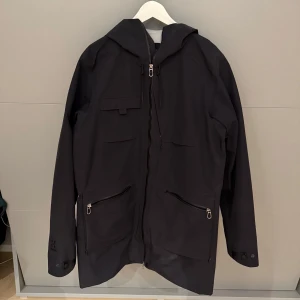 Svart vindjacka från Haglöfs - Säljer en svart vindjacka från Haglöfs i storlek M. Jackan har en praktisk huva och dragkedja framtill. Perfekt för blåsiga dagar och har en stilren design med två fickor med dragkedjor. Passar bra för både vardag och utomhusaktiviteter.