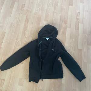 Säljer en snygg svart hoodie från Lacoste i storlek L. Den har en dragkedja framtill och den klassiska krokodilloggan på bröstet. Perfekt för höst och vinter, och den är superbekväm att ha på sig. Passar både till vardags och chillkvällar. Hör av dig om du är intresserad!