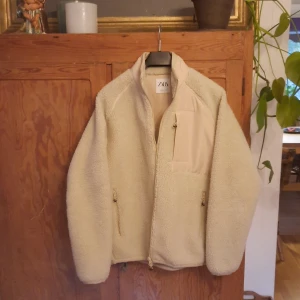 Beige teddyjacka från Zara - Säljer en supermysig beige teddyjacka från Zara i storlek M. Jackan har en dragkedja framtill och en praktisk bröstficka. Perfekt för kyliga höst- och vinterdagar när du vill hålla dig varm och stilren. Den är i nyskick och redo för nya äventyr!