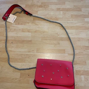 Röd väska - En liten röd handväska med silver kedja och stjärnor på framsidan. Aldrig använd och lappen sitter kvar. Den är 16,5 cm bred och 14 cm hög och stängs med magnet.