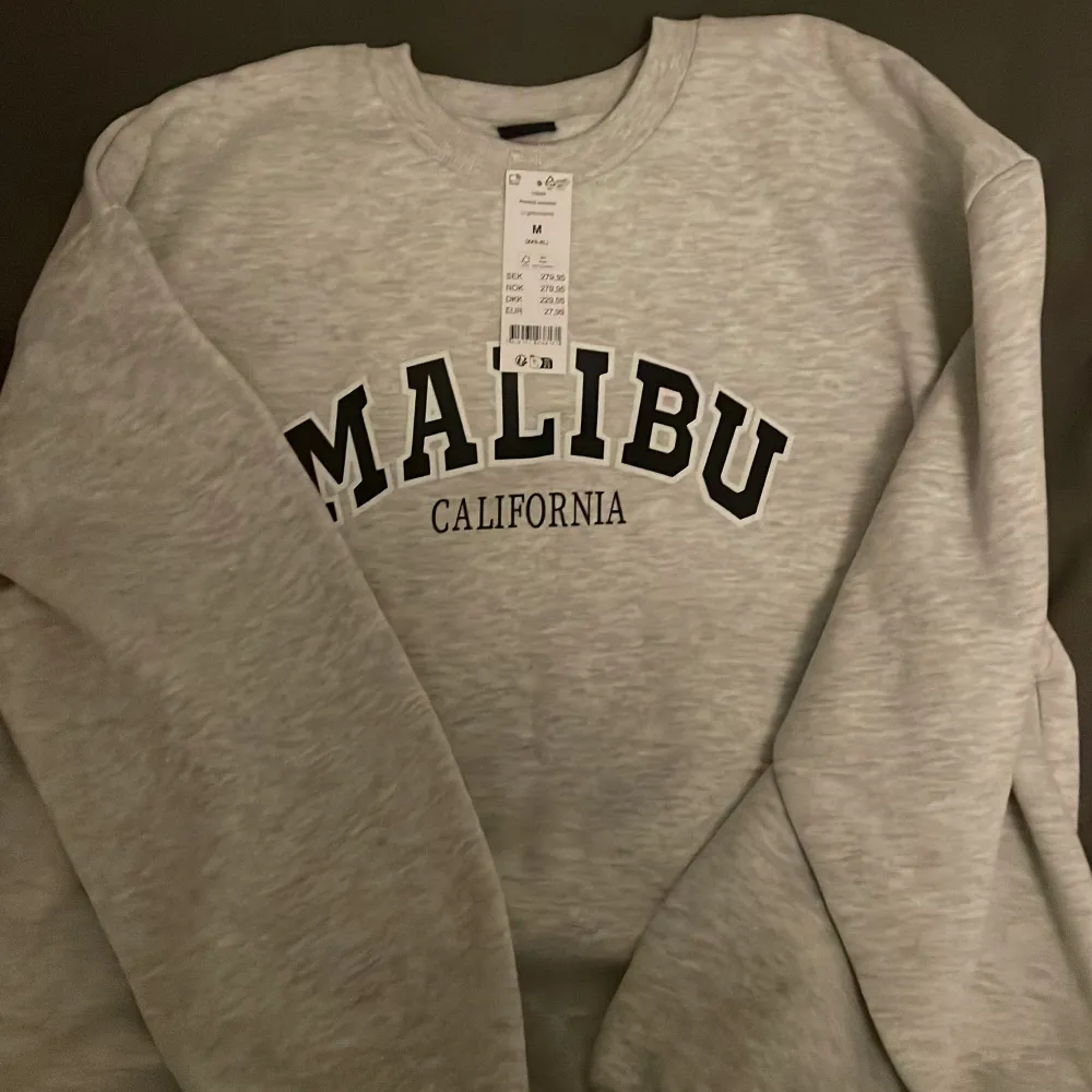 Säljer en supermjuk och bekväm grå sweatshirt från Gina med texten 'Malibu California' tryckt i svart på framsidan. Perfekt för en avslappnad stil och passar till alla tillfällen. Den har långa ärmar och en klassisk rund halsringning. Helt ny med t. Tröjor & Koftor.