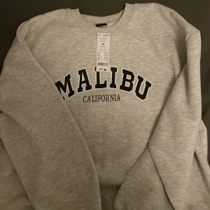 Grå sweatshirt med Malibu-tryck - Säljer en supermjuk och bekväm grå sweatshirt från Gina med texten 'Malibu California' tryckt i svart på framsidan. Perfekt för en avslappnad stil och passar till alla tillfällen. Den har långa ärmar och en klassisk rund halsringning. Helt ny med t