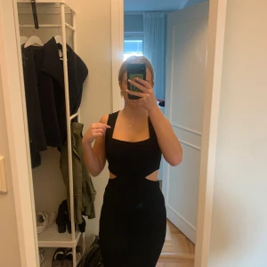 Svart klänning med cut-out detaljer - Säljer en elegant svart klänning med trendiga cut-out detaljer i midjan. Klänningen är ärmlös och har en tight passform som framhäver figuren. Perfekt för festliga tillfällen eller en utekväll. Den är gjord i ett mjukt och stretchigt material som ger en bekväm känsla hela kvällen.