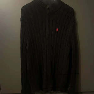 Svart stickad tröja från Ralph Lauren - Säljer en snygg svart stickad tröja från Ralph Lauren. Den har en dragkedja vid halsen och det klassiska röda logotypen på bröstet. Perfekt för kyliga dagar och ger en stilren look. Passar både till vardags och lite finare tillfällen.
