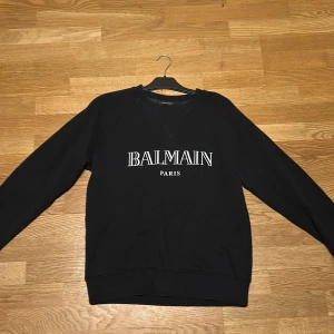 Svart sweatshirt från Balmain - Säljer en stilren svart sweatshirt från Balmain med deras ikoniska logga tryckt på framsidan. Tröjan är tillverkad i mjuk bomull och har långa ärmar samt ribbade muddar vid ärmslut och nederkant. Perfekt för en casual look.  Skick: 9/10 (använd fåtal gånger). Priset går att diskutera!