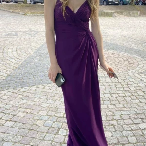 Mörklila balklänning från JJ's House - Elegant mörklila långklänning med v-ringning och draperad detalj framtill. Klänningen har en öppen rygg med korsade band som ger en snygg look. Perfekt för speciella tillfällen.