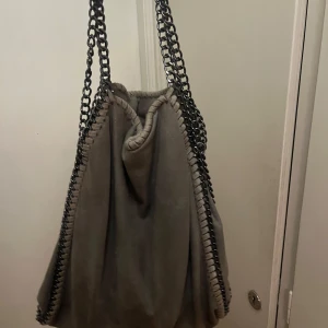 Grå axelväska med kedjedetaljer - Snygg grå axelväska med coola kedjedetaljer längs kanterna. Väskan har en mjuk och slouchy design, perfekt för en avslappnad stil. Den är rymlig och passar bra för att bära med sig allt man behöver under dagen.