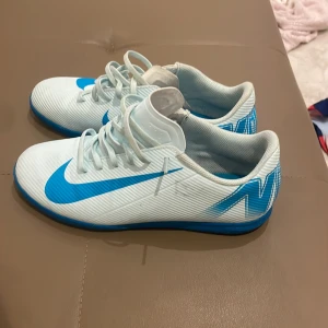 Vita och blå fotbollsskor från Nike - Snygga vita fotbollsskor från Nike med blå detaljer och snörning. Perfekta för inomhusspel med en stilren design och bra grepp på sulan.