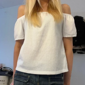 Vit offshoulder topp med spetsdetaljer - Säljer en söt vit offshoulder topp med spetsdetaljer på baksidan. Toppen har korta ärmar och en luftig passform, perfekt för varma dagar. Den är stilren och lätt att matcha med olika outfits.