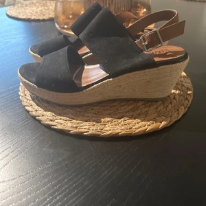 Svarta sandaler med kilklack - Snygga svarta sandaler från Linea med kilklack och justerbart spänne. Ovandelen är i mocka och sulan har en flätad design som ger en somrig känsla. Perfekta för varma dagar.
