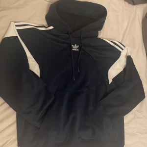 Mörkblå hoodie från Adidas - Säljer en snygg mörkblå hoodie från Adidas med vita detaljer och klassiska ränder på axlarna. Tröjan har en stor luva och en praktisk magficka framtill. Perfekt för en sportig look eller en avslappnad dag.