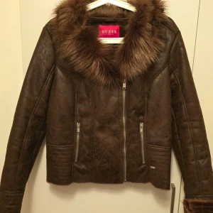 Brun skinnjacka med fuskpäls från Guess - Snygg brun skinnjacka från Guess med fuskpäls vid kragen. Jackan har dragkedja framtill och två fickor med dragkedjor. Perfekt för kyligare dagar med sin stilrena design och varma foder.