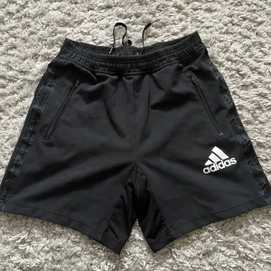 Svarta shorts från Adidas - Snygga svarta shorts från Adidas med dragkedja på fickorna och elastisk midja med snörning. Adidas-loggan är tryckt på framsidan och det finns ett subtilt mönster längs sidorna. Perfekta för träning eller en avslappnad dag.