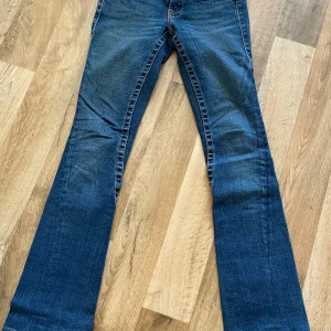 Blå bootcut jeans från True Religion - Säljer ett par snygga blå bootcut jeans från True Religion. De har en klassisk femficksdesign och märkets ikoniska logga på bakfickorna. Perfekta för dig som gillar en tidlös stil med en modern twist. 
