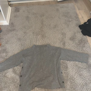 Grå tröja med knappar - Säljer en stilren grå tröja med knappar på sidorna. Tröjan har långa ärmar och en enkel design som passar perfekt för en avslappnad look. Perfekt för kyligare dagar!