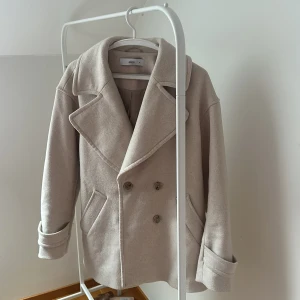 Beige kappa från Ellos - Säljer en stilren beige kappa från Ellos i storlek 36. Den har en klassisk dubbelknäppt design med breda slag och långa ärmar. Perfekt för höst och vår. Kappan har en mjuk och bekväm känsla.