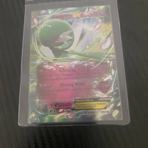 Gardevoir EX Pokémonkort - Säljer ett Gardevoir EX Pokémonkort med 170 HP. Kortet har attacker som Life Leap och Shining Wind. Det är ett holografiskt kort med en färgglad design och är skyddat i en plastficka.