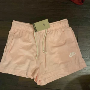 Rosa shorts från Nike - Säljer ett par snygga rosa shorts från Nike med elastisk midja och dragsko. Perfekta för en avslappnad stil. Nike-loggan i vitt på benet ger en sportig touch. Passar perfekt för varma dagar.
