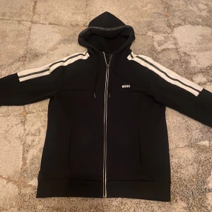 Svart zip med dragkedja från Boss - Snygg svart jacka från Boss med dragkedja och vita detaljer längs ärmarna. Perfekt för en stilren look. Jackan har långa ärmar och en klassisk passform, hej säljer en helt ny Hugo boss kofta den är knappt använd och köpt för 2200kr tveka inte på prisförslag går ner i pris vid snabb affär