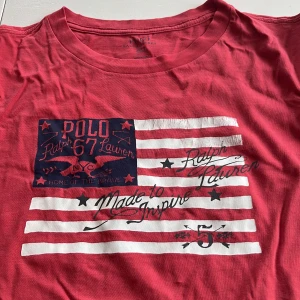Röd t-shirt från Ralph Lauren - Säljer en röd t-shirt från Ralph Lauren med ett tryck av amerikanska flaggan och texten 'Polo 67 Ralph Lauren'. Perfekt för en avslappnad stil. Tillverkad i mjuk bomull.