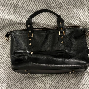 Svart skinnväska med dragkedja - Snygg svart skinnväska med guldfärgade detaljer och dragkedja. Väskan har två handtag och en röd insida som ger en fin kontrast. Perfekt för att bära dina dagliga nödvändigheter med stil.