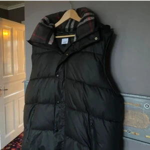 Burberry Väst/Jacka - Säljer en riktigt snygg o trendig svart dunjacka från Burberry med avtagbara ärmar, vilket gör den till en praktisk väst. Perfekt för kommande väder! Jackan har en quiltad design och en huva med rutigt mönster på insidan. Perfekt för kyliga dagar o mindre kyliga! Har en liten skada på dragkedjan. (Se sista bilden) Men den fungerar fortfarande👍💯