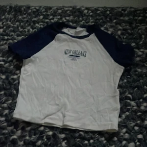 Vit och marinblå kortärmad tröja  - Säljer en cool vit t-shirt med marinblå ärmar från H&M. Tryck med texten 'New Orleans Gulf Coast' på framsidan. Perfekt för en avslappnad stil.