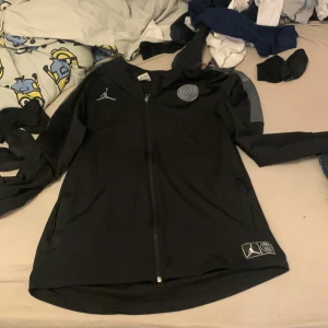 Svart jacka från Jordan x PSG - Säljer en svart jacka från Jordan i samarbete med Paris Saint-Germain. Jackan har en dragkedja framtill och är dekorerad med PSG-loggan och Jumpman-märket. Perfekt för fotbollsfans och streetwear-entusiaster.