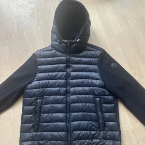 Mörkblå Colmar Cardigan - Snygg mörkblå Cardigan  med huva och dragkedja framtill. Jackan har långa ärmar och är quiltad på framsidan med slät baksida. Perfekt för våren 