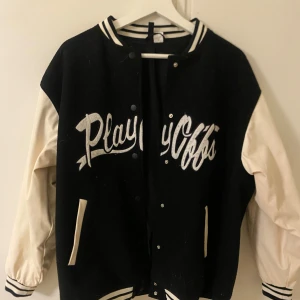 Svart och vit bomberjacka med text - Snygg svart och vit bomberjacka med texten 'Playoffs' på framsidan och siffran '92' på ärmen. Jackan har långa ärmar och stängs med knappar. Den har ribbade muddar och krage i svart och vitt.