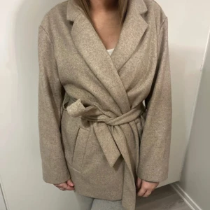Beige kappa från ONLY - Säljer en stilren beige kappa från ONLY i storlek S. Den har en klassisk omlott-design med ett knytbälte i midjan för en snygg passform. Perfekt för höst och vår med sina långa ärmar och mjuka material. Nypris 600 men säljer för 200