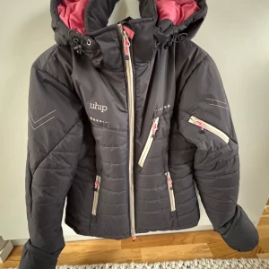 Grå dunjacka från Uhip - Säljer en grå dunjacka från Uhip med rosa foder och flera praktiska fickor med dragkedja. Jackan har en justerbar huva och är perfekt för kyliga dagar. Den har en snygg design med diskreta detaljer och en bekväm passform. Nyskick!!