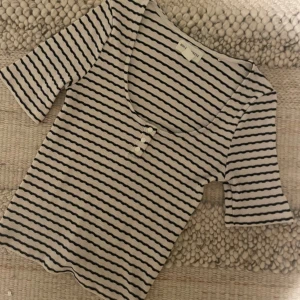 Randig topp från Holly & Whyte - Säljer en randig topp från Holly & Whyte by Lindex i svart och vit. Toppen har en rund halsringning och trekvartsärmar med knappar framtill. Perfekt för en stilren look.