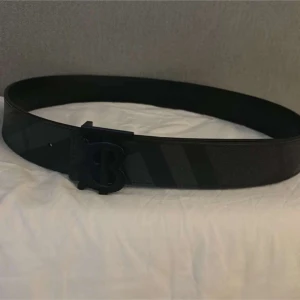 Svart bälte med monogramspänne - Snyggt svart bälte med ett stilrent monogramspänne. Bältet har en diskret randig design som ger det en modern touch. Perfekt för att lyfta vilken outfit som helst.