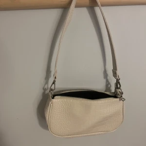 Beige axelväska med krokodilmönster - Snygg beige axelväska med krokodilmönster och dragkedja. Perfekt för att ge din outfit en trendig touch. Väskan har en justerbar axelrem och är tillverkad i syntetmaterial. Superbra skick då den bara används en gång. Pris kan diskuteras!❤️