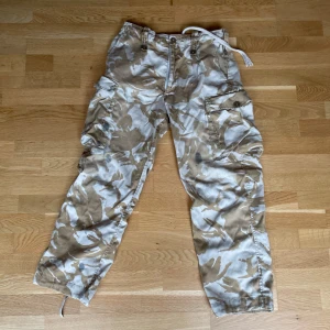 NATO kamouflage cargobyxor - Säljer ett par kamouflagefärgade cargobyxor i beige och brunt. Äkta från NATO. Oljefläckar lite här och där. Snörena är lite sönder men fungerar fortfarande.