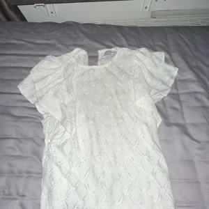 Vit topp från Holly & Whyte - Säljer en vit topp från Holly & Whyte med ett diskret mönster. Toppen har korta ärmar och en rund halsringning. Perfekt för en stilren look.