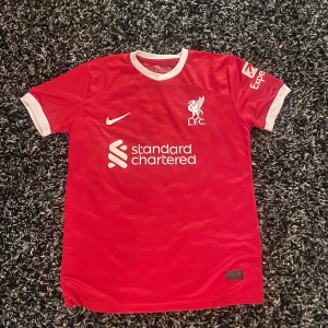Röd Liverpool fotbollströja - Säljer en röd Liverpool fotbollströja från Nike med vit krage och ärmslut. Tröjan har klubbens emblem och sponsorlogga på framsidan. Perfekt för alla LFC-fans! 🔴