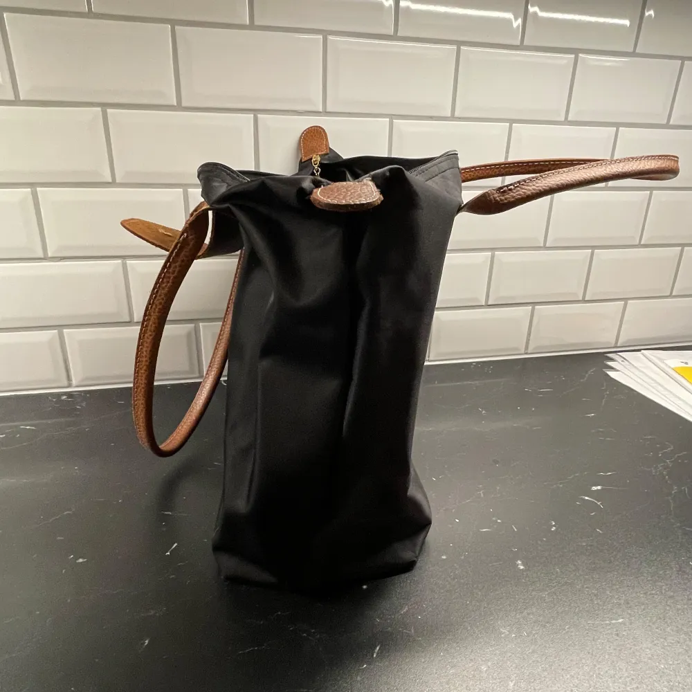 Snygg svart väska som LIKNAR en från Longchamp med bruna skinnhandtag och en praktisk dragkedja. Väskan har en klassisk design med en knappdetalj framtill och är perfekt för att bära med sig det nödvändigaste. En stilren accessoar för alla tillfällen.. Laukut & Käsilaukut.
