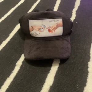 Svart keps från Lyxery - Snygg svart keps från Lyxery med ett unikt tryck framtill som visar två händer med texten 'First Class Statement' och 'No Risk No Story'. Kepsen har ett nätmaterial baktill för ventilation och en justerbar rem för passform.