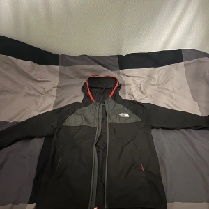Svart jacka från The North Face - Säljer en svart jacka från The North Face med luva och dragkedja. Jackan har röda detaljer och logotypen tryckt på bröstet och ryggen. Perfekt för varmare dagar.