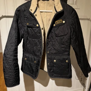 Svart quiltad jacka från Barbour - Säljer en snygg svart quiltad jacka från Barbour med mjukt foder. Jackan har knappar och dragkedja framtill samt flera praktiska fickor. Perfekt för kyligare dagar.