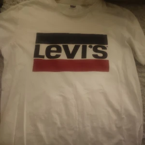 Vit t-shirt från Levi's - Säljer en klassisk vit t-shirt från Levi's med det ikoniska logotyptrycket i svart och rött på bröstet. Perfekt för en avslappnad stil.
