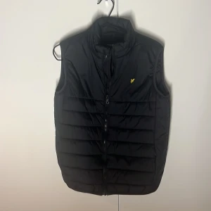 Svart dunväst från Lyle & Scott - Snygg svart dunväst från Lyle & Scott med dragkedja och en liten gul logga på bröstet. Perfekt för kyligare dagar med sin quiltade design och höga krage. Köpt för 799 kr