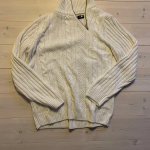 Vit stickad tröja från JC - Säljer en stilren vit stickad tröja från JC med dragkedja vid halsen. Tröjan har ett klassiskt flätmönster och långa ärmar, perfekt för kyligare dagar. Den ribbade kragen ger extra värme och stil. Passar utmärkt till både jeans och chinos.