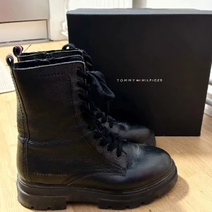 Svarta kängor från Tommy Hilfiger - Snygga svarta boots i 100% läder från Tommy Hilfiger med snörning och dragkedja på insidan. De har en robust sula 5,5 cm och klassisk design med märkets logotyp diskret placerad. Skafthöjd 18 cm. Använda 2 ggr och i mycket fint skick! Perfekta för en stilren look.