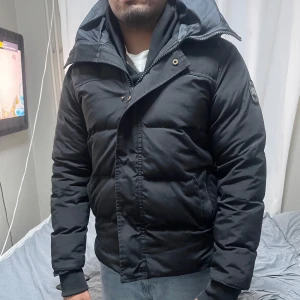 Black label Canada Goose pufferjacket - Säljer en svart black label jacka från Canada Goose. Jackan har en pufferdesign med långärmade ärmar och en praktisk dragkedja framtill. Perfekt för kalla dagar med sin varma och isolerande fyllning. Märkeslogga på ärmen ger en stilren touch.